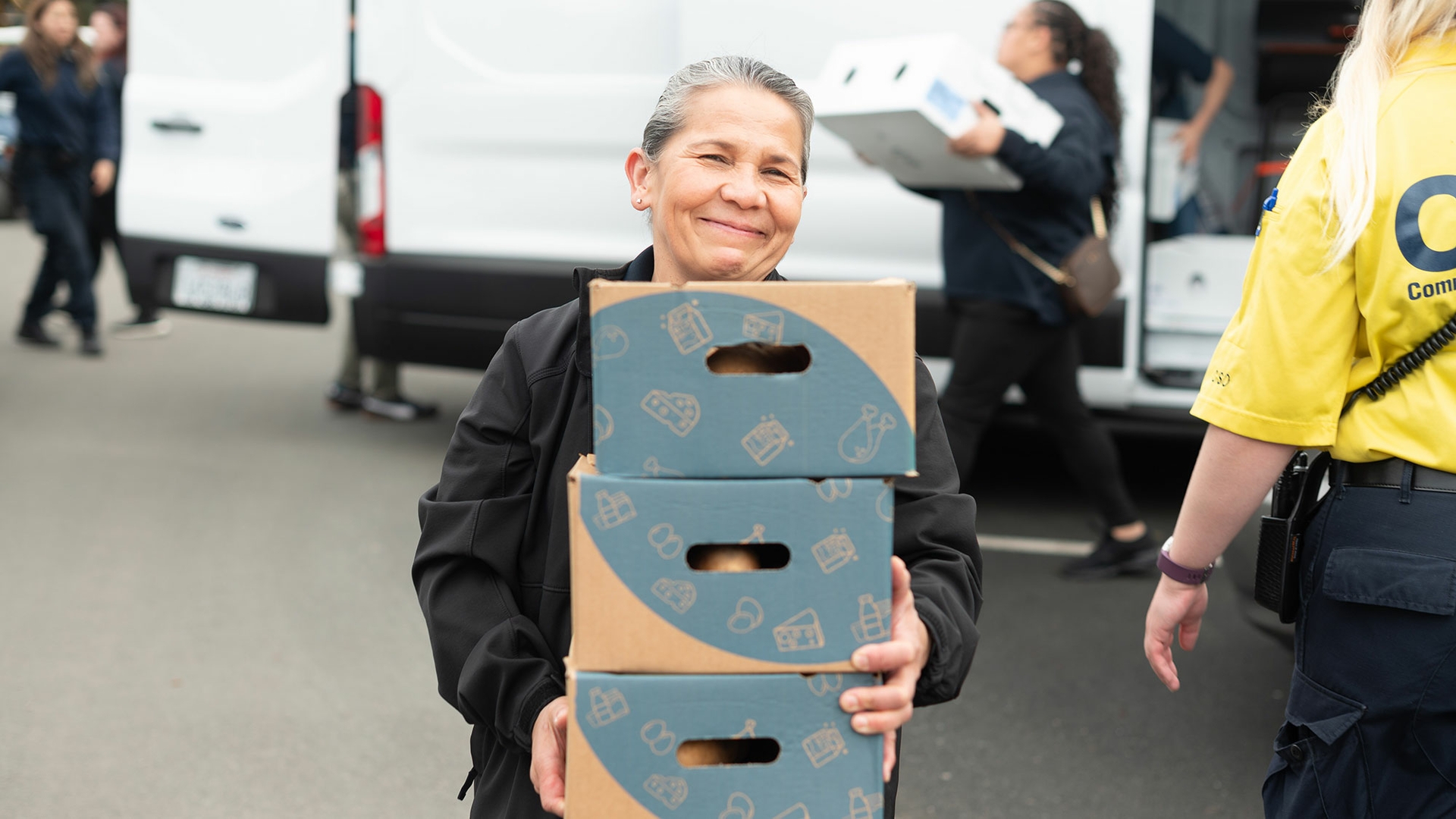 A volunteer wearing a black jacket smiles at the camera while carrying a stack of three cardboard boxes.