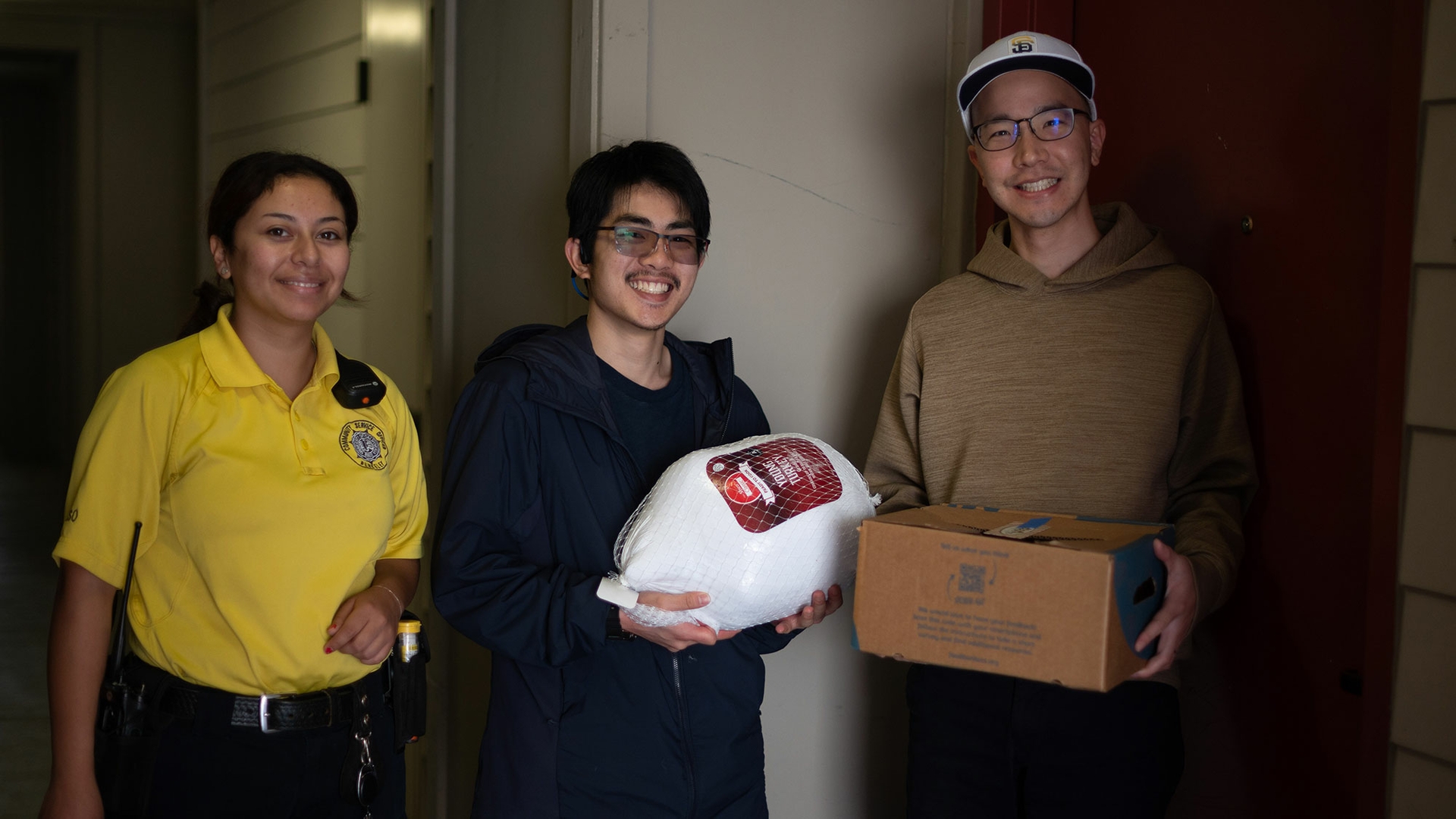 Three people stand beside the entryway to a residence. The person in the middle is holding a large frozen turkey, and the person next to them is holding a cardboard box.