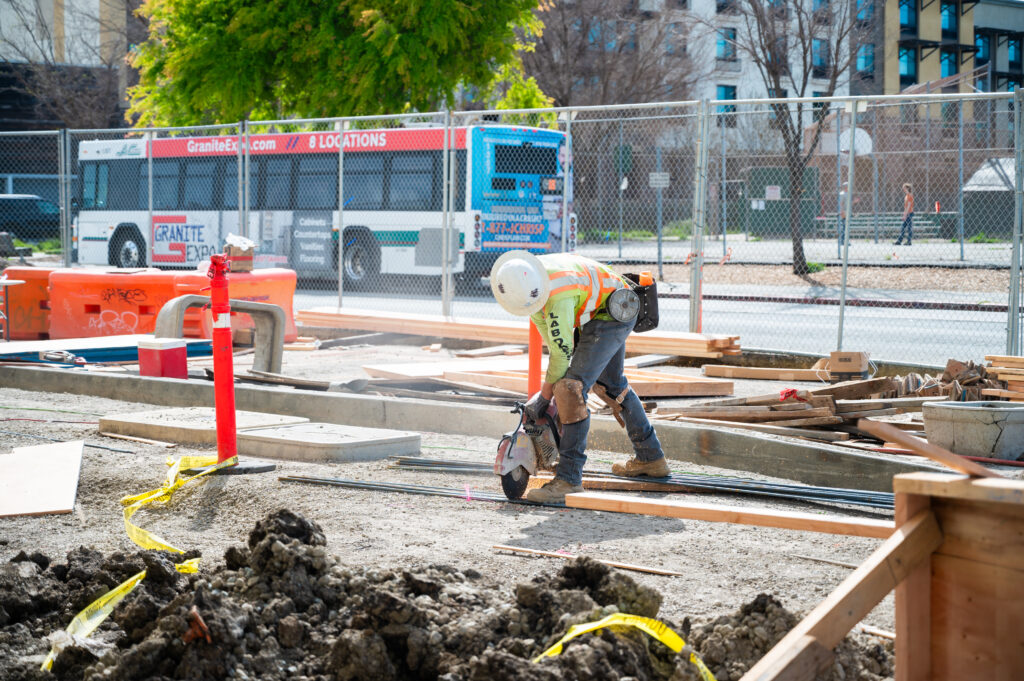 A worker wearing a construction helmet, kneepads and a safety vest saws wood outside the new graduate student apartments in Albany. He is helping to construct a transit stop for AC Transit buses.