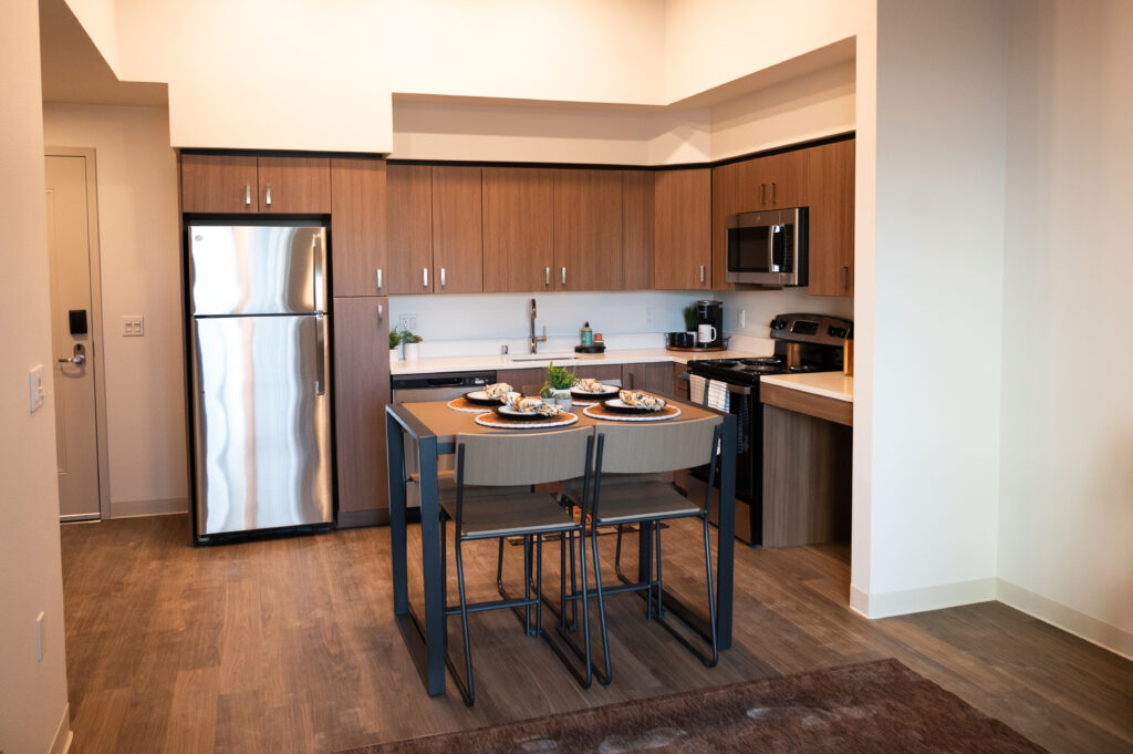 A view of the kitchen and eating area in the new graduate student apartments opening in Albany in fall 2024. There is wood-look flooring, stainless steel appliances and a tall table with bar stool-height chairs.