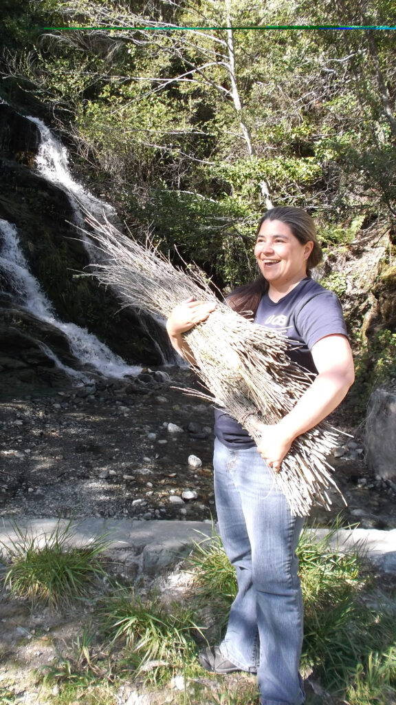 Carolyn standing by a waterfall wraps her arms around a bundle of willow sticks