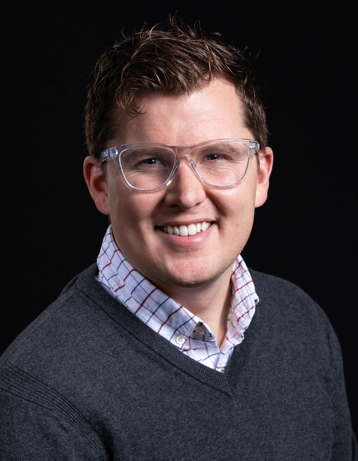 Headshot of Andrew Reddie, wearing a grey v-neck sweater over a plaid shirt, against a dark background