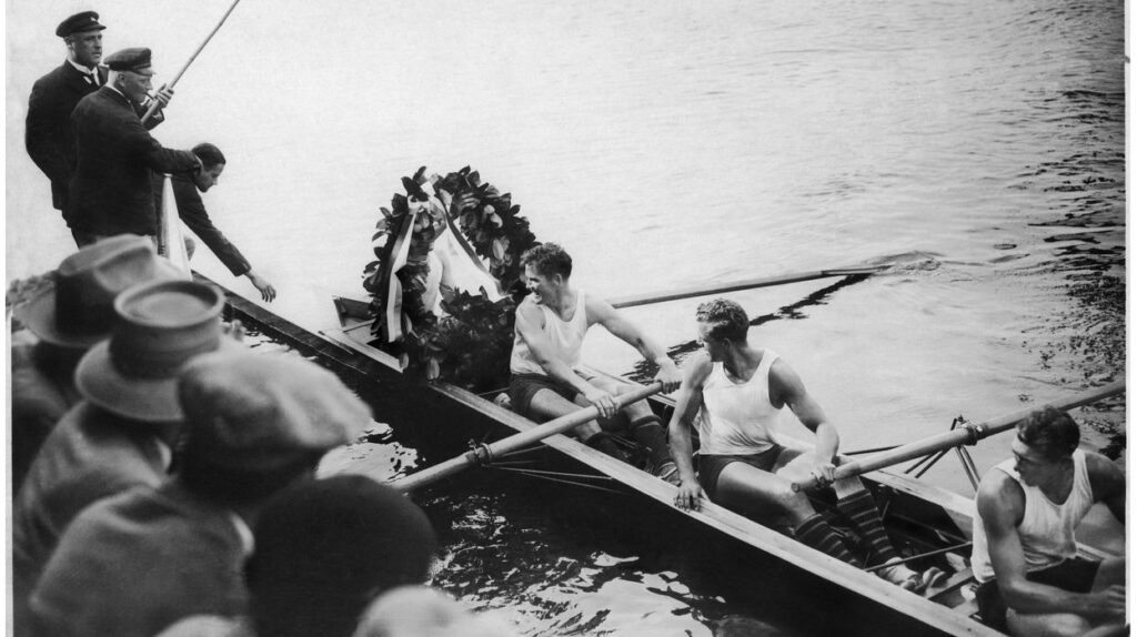 a black and white image of a narrow boat near a dock with men inside rowing"