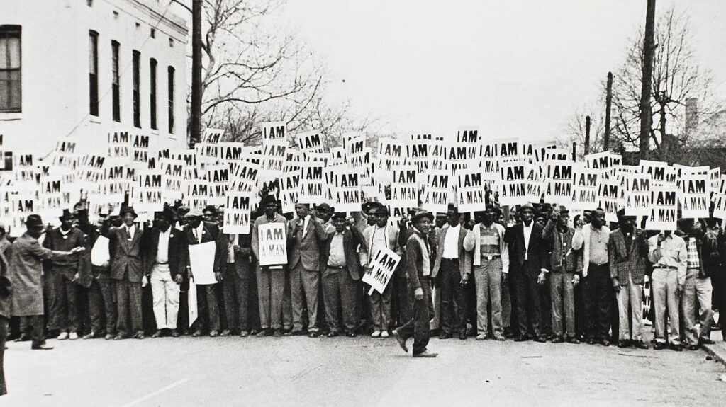 At a 1957 civil rights protest, scores of Black sanitation workers in a black & white photo carry placards reading “I Am a Man”