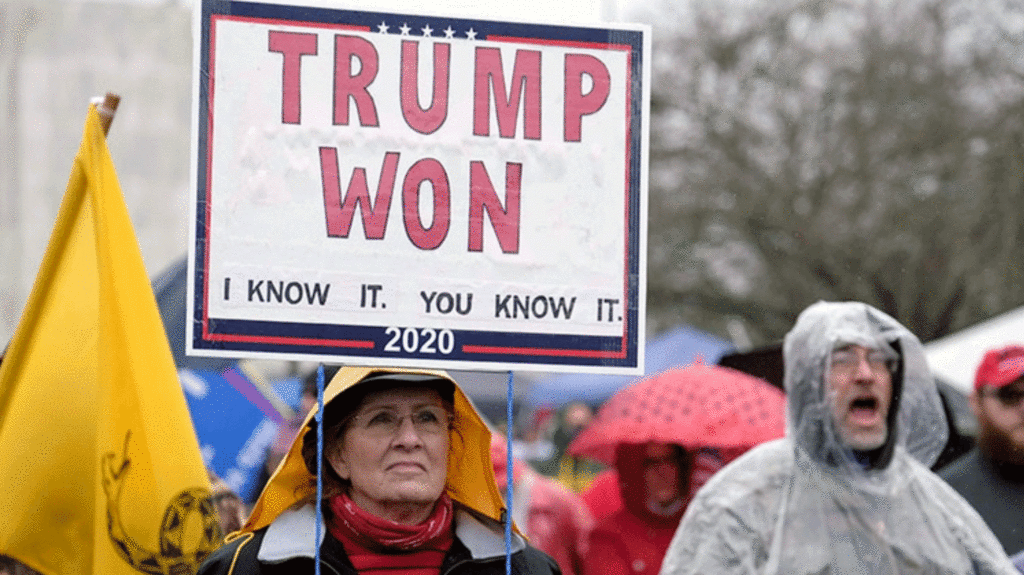 On a rainy day soon after the 2020 presidential election, a woman standing with other protesters holds a sign saying: