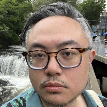 Headshot of a person with short gray and black hair wearing tortoiseshell glasses, standing in front of a water fall.