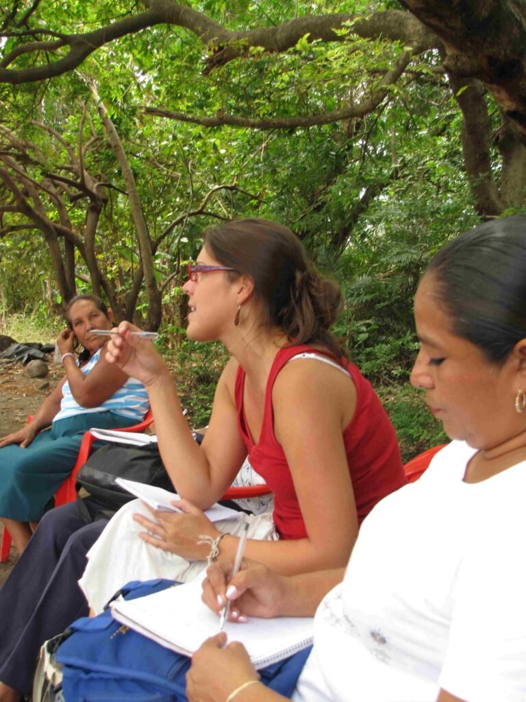 Tedde Simon, the campus's new tribal liaison sits in a circle of women in El Salvador in 2010, where she is facilitating a discussion. The women are sitting under leafy green trees and holding notebooks and pens.