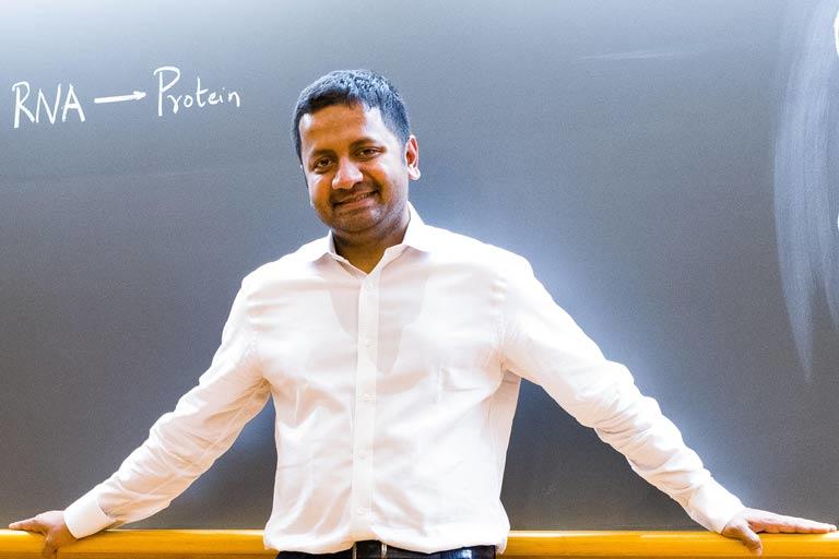 man in white shirt standing in front of chalkboard with the words 'RNA to Protein'