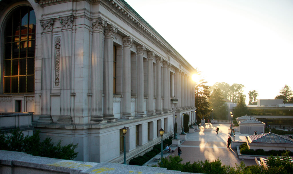 A photo of Doe Library with the sun peeking out around a far corner of the building's exterior.