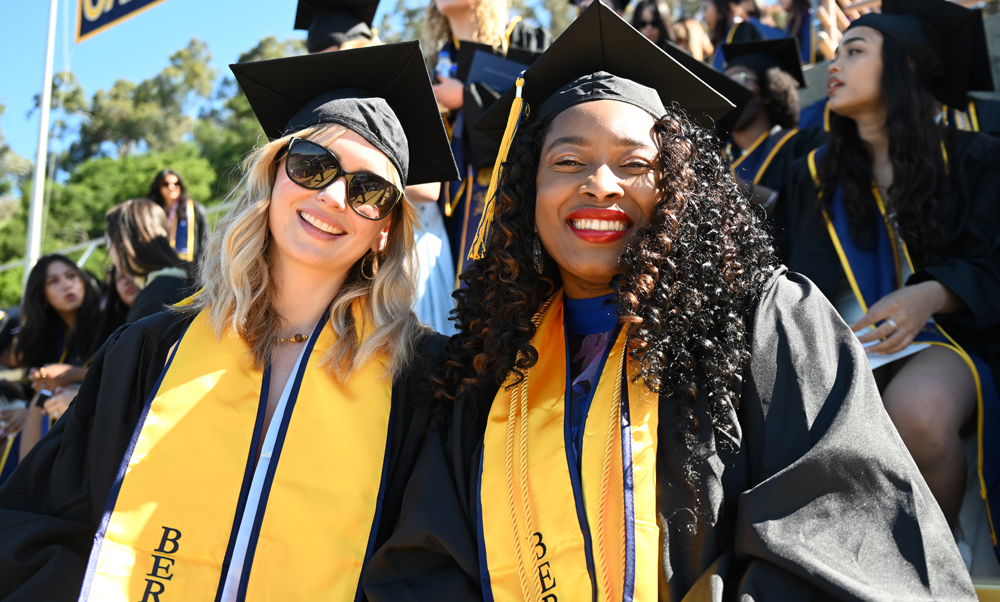 Two women smiling, wearing mortarboards and gowns.