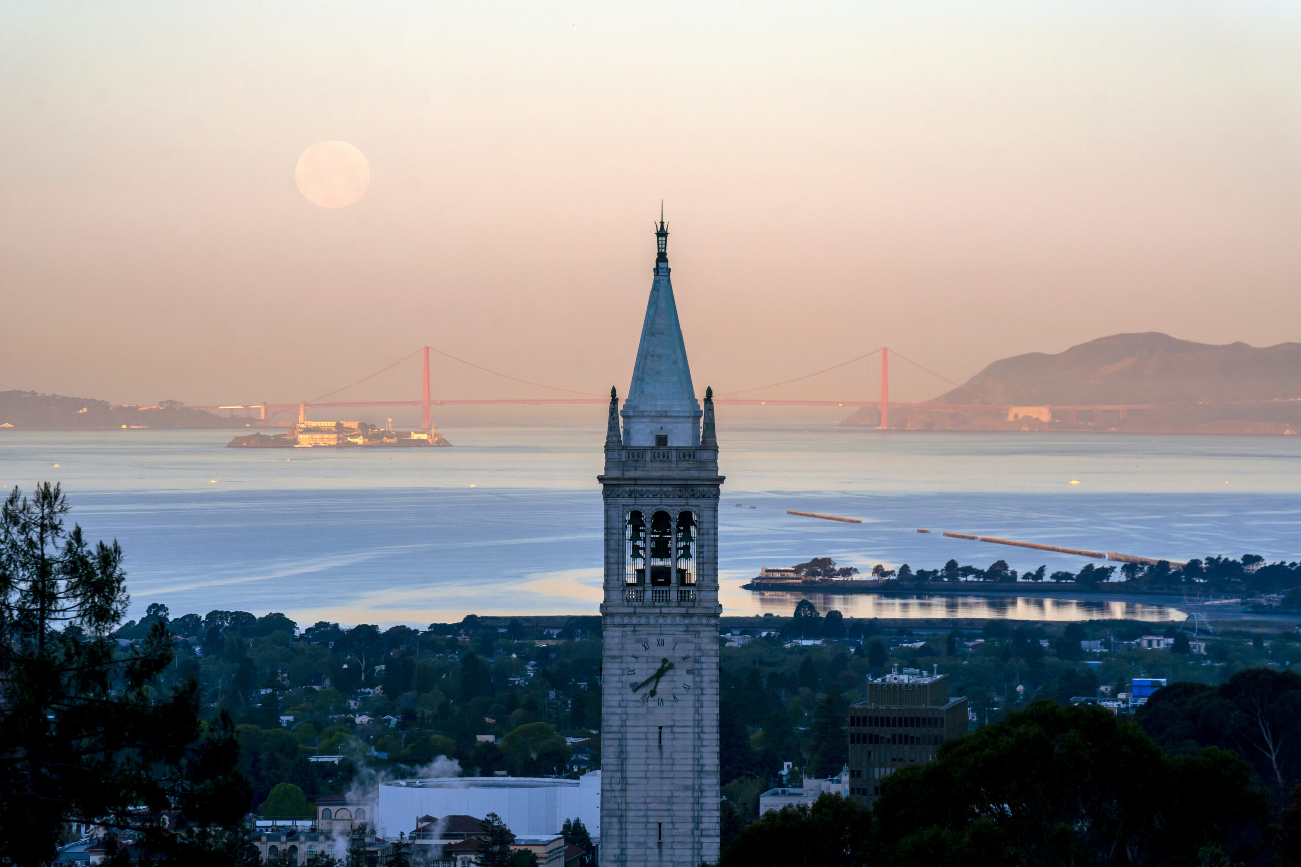 The Campanile in the evening, with a pinkish sky and a supermoon in the background, where you can also see the Golden Gate Bridge.