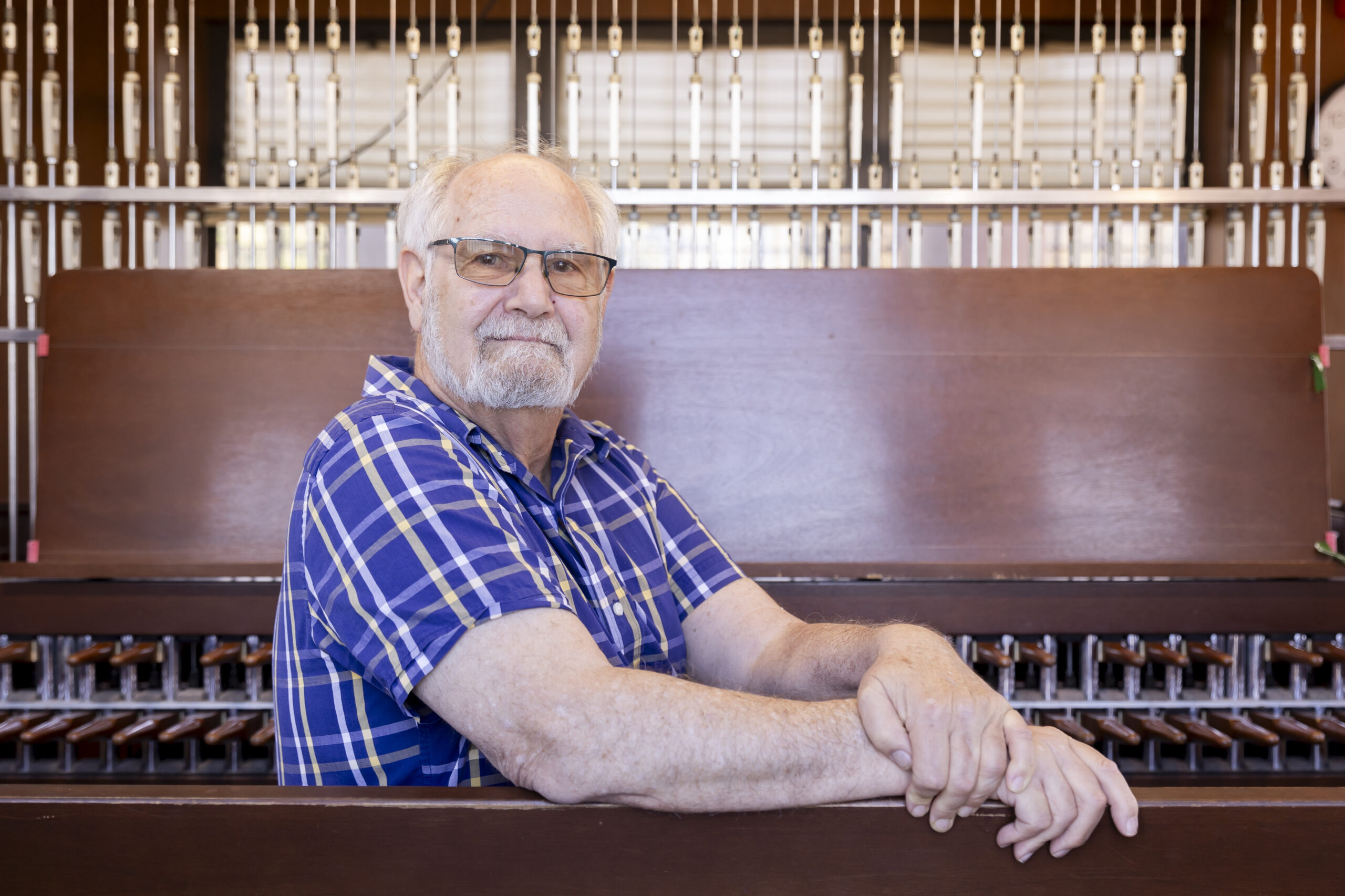 Jeff Davis, the retiring university carillonist, sits on the bench of the grand carillon on the observation deck of the Campanile. His body is turned toward the camera and he's wearing eye glasses, a beard, a short-sleeved plaid shirt and a smile.