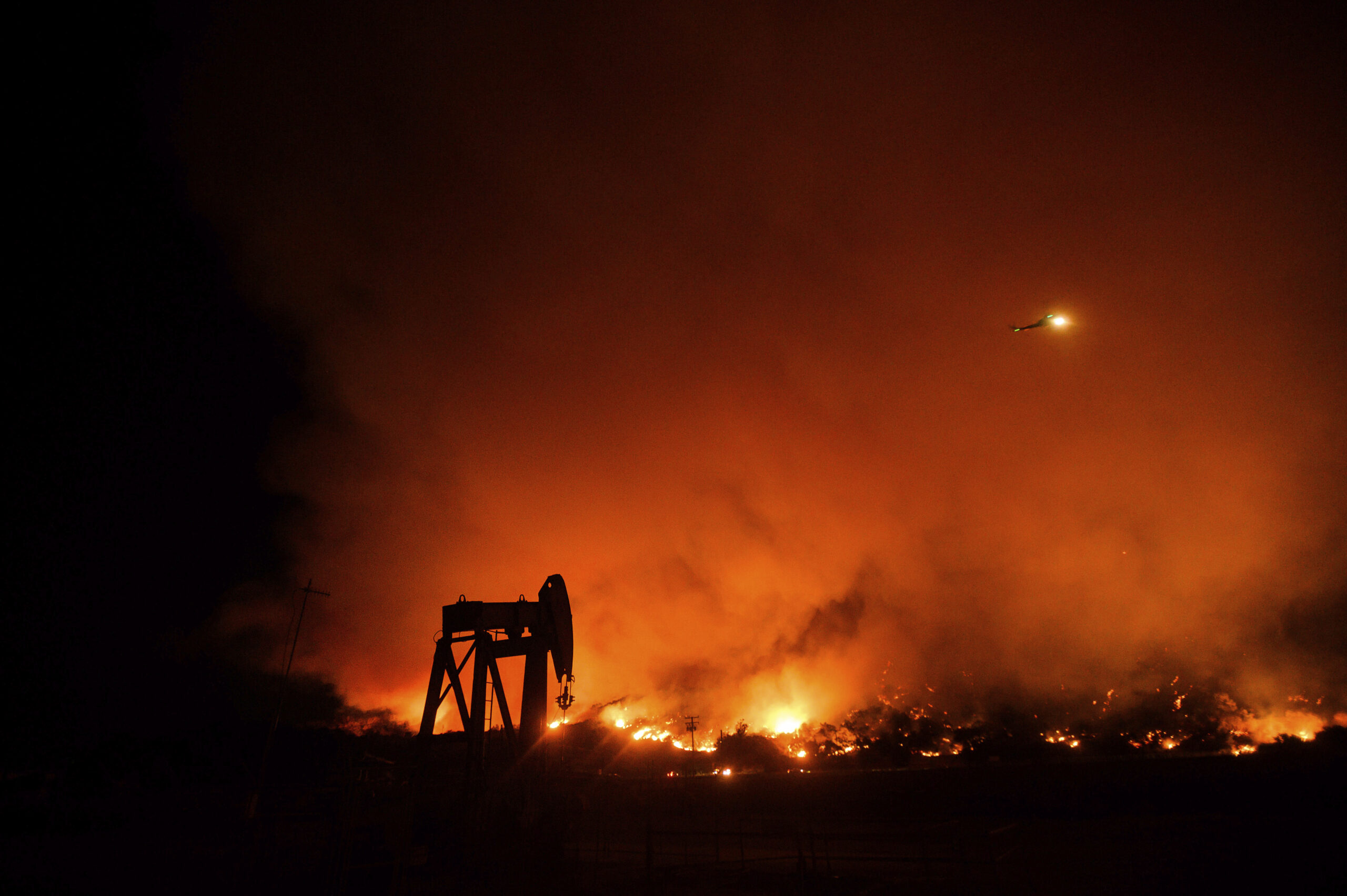 orange flames from a wildfire in California burning at night in the distance, with the silhouette of an oil and gas pumpjack in the foreground.
