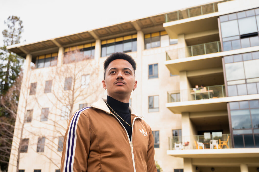 William Carter stands outside the building that houses the geography department on campus, wearing a tan-colored Adidas track suit