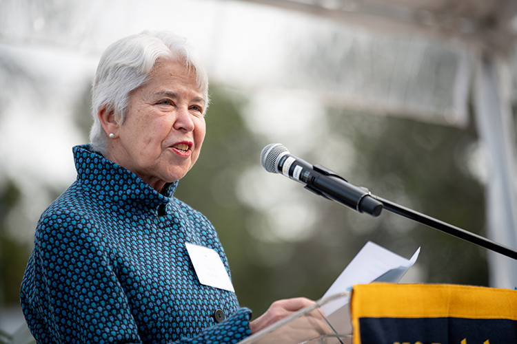 Chancellor Carol Christ speaks at a microphone wearing a printed coat in shades of blue. She is speaking at the groundbreaking ceremony for Anchor House.