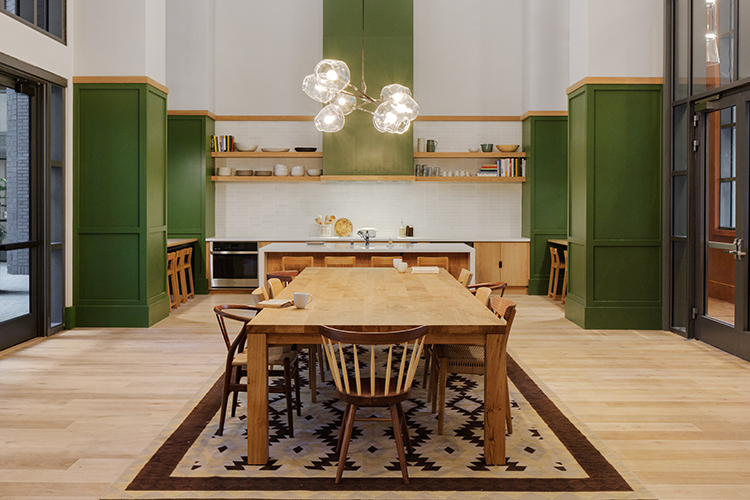 An area of Anchor House called The Pantry is a beautiful, fully-equipped kitchen with cabinets and an oven hood painted olive green. A large wooden table with unique wooden chairs is a place for residents to share a meal. There is a hardwood floor and open shelving above the countertops.