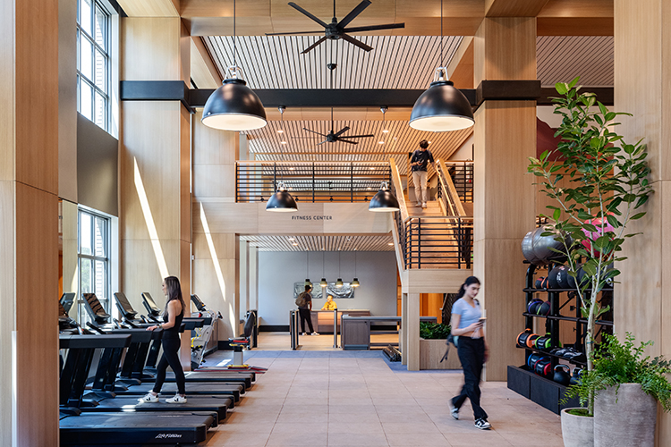 Anchor House has an 8,600-square-foot space that contains a fitness center and yoga studio. A student is using a treadmill, while another is walking down a staircase to the fitness center from the yoga studio above. There are ceiling fans and large windows that light is shining through.