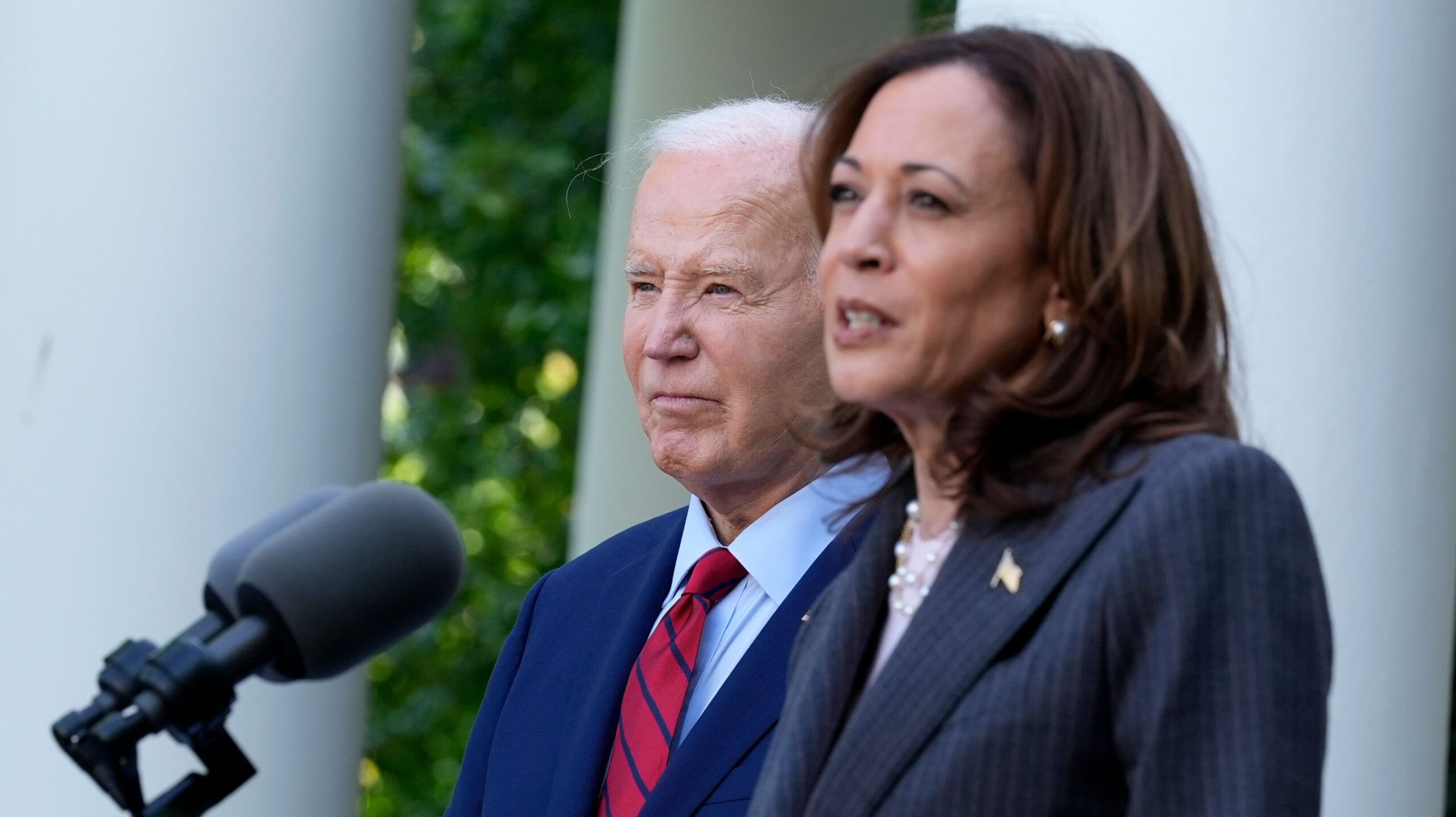 Vice President Kamala Harris speaks into a microphone outside of the White House, with a serious expression. President Joe Biden is to her side and slightly behind her, also looking serious.
