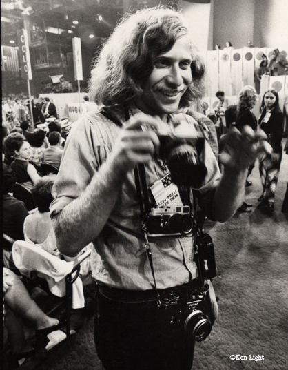 A black-and-white photo from 1972 shows professor Ken Light as a young photojournalist holding a camera at the Republican National Convention in Miami. He has long frizzy hair and is smiling.