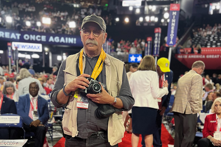 Ken LIght, a Berkeley professor and documentary photojournalist, stands on the red carpeting of the Republican National Convention in Milwaukee holding a camera and wearing a cap and vest.