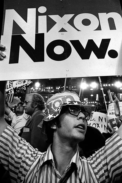 A man in a short-sleeved striped shirt holds up a "Nixon Now." poster during a demonstration outside of the 1972 Republican National Convention in Miami.