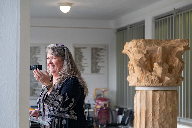 Kim Shelton, a professor of Ancient Greek and Roman studies on campus, stands next to an ancient column at an exhibit at the campus's Nemea Center for Classical Archaeology in Ancient Nemea, Greece. She is gesturing to the audience with her left hand while speaking at a microphone.