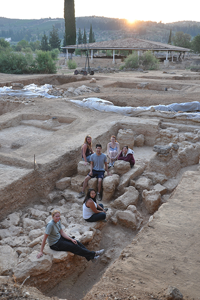 Students sit on the ground or stand within an archaeological excavation area where they've been working in Ancient Nemea, Greece, where the Panhellenic Games were once held.