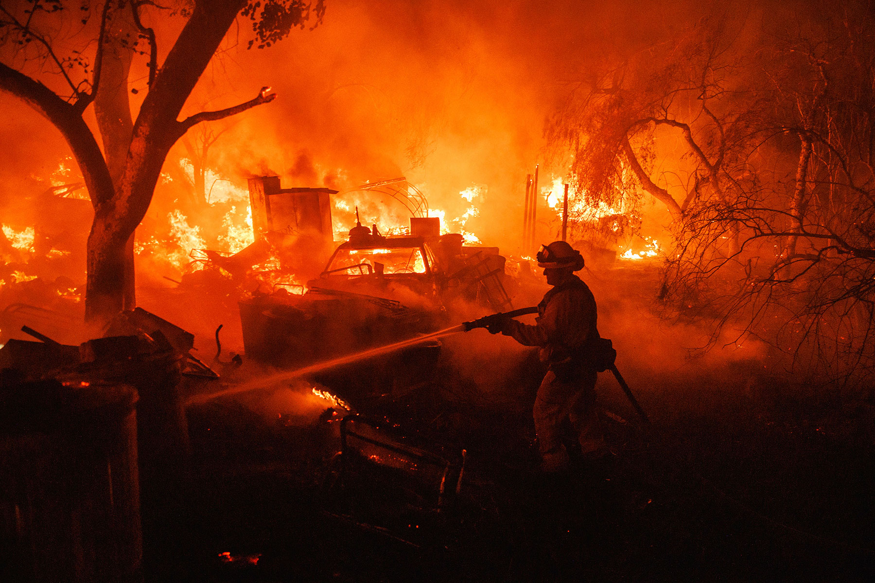 A firefighter points a hose at a burning structure, while orange flames blaze in the background.
