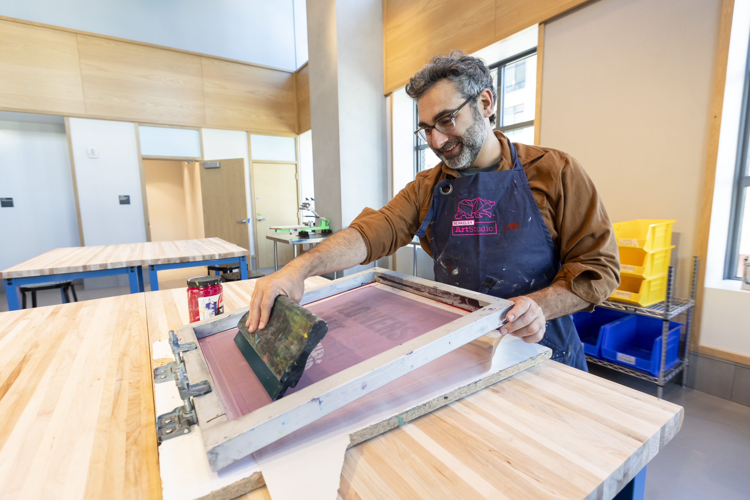 A printmaker practices his craft with a smile at Berkeley Art Studio West, the new satellite facility for Berkeley Art Studio in the MLK Jr. Building. He has a beard, mustache and eyeglasses and is working on a butcher block table with his printmaking equipment.