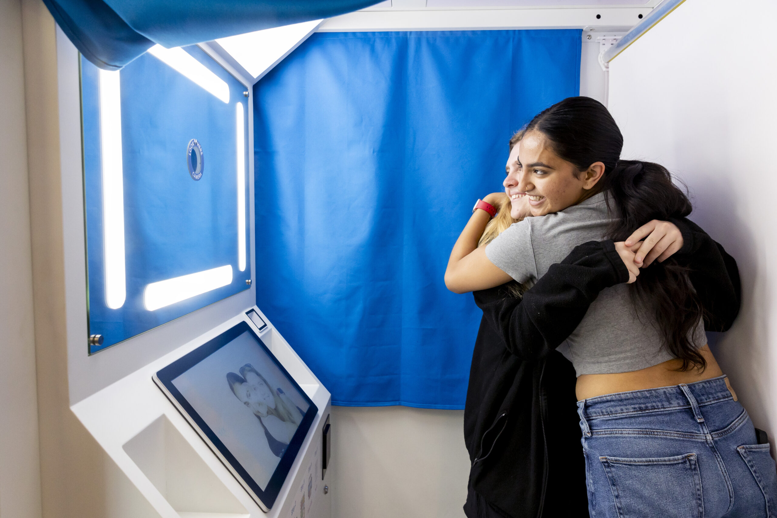 Two students hug each other and smile inside a new photo booth in the Martin Luther King Jr. Buildling of the student union. A blue curtain is pulled behind them, and they can see their image on a screen at waist level in front of them.