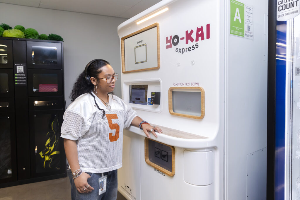 A student stands at a new robot in the MLK Jr. Building of the student union that dispenses ramen noodles. The white machine is much taller than the student and says Yo-Kai Express on it. There is a digital screen and a place to scan your payment card, then a window that says Caution Hot Bowl above the little door that opens with the student's order.