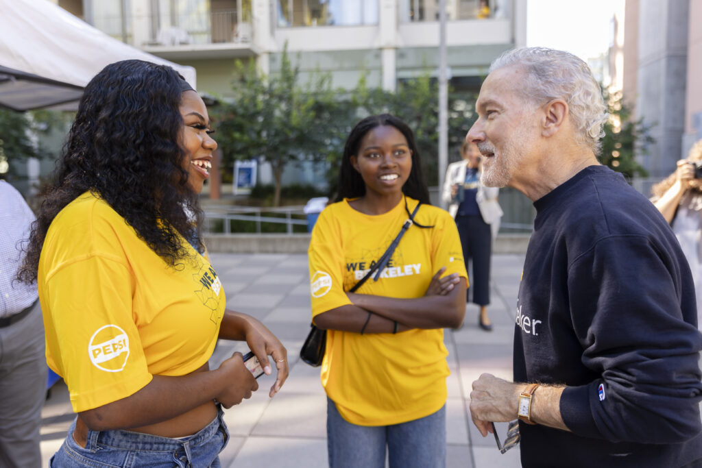 Chancellor Lyons, wearing a Berkeley Changemaker sweatshirt, talks with a campus RA, a resident assistant, during one of the fall move-in days. She is wearing a gold "We Are Berkeley" T-shirt and jeans and is smiling while speaking with Lyons.