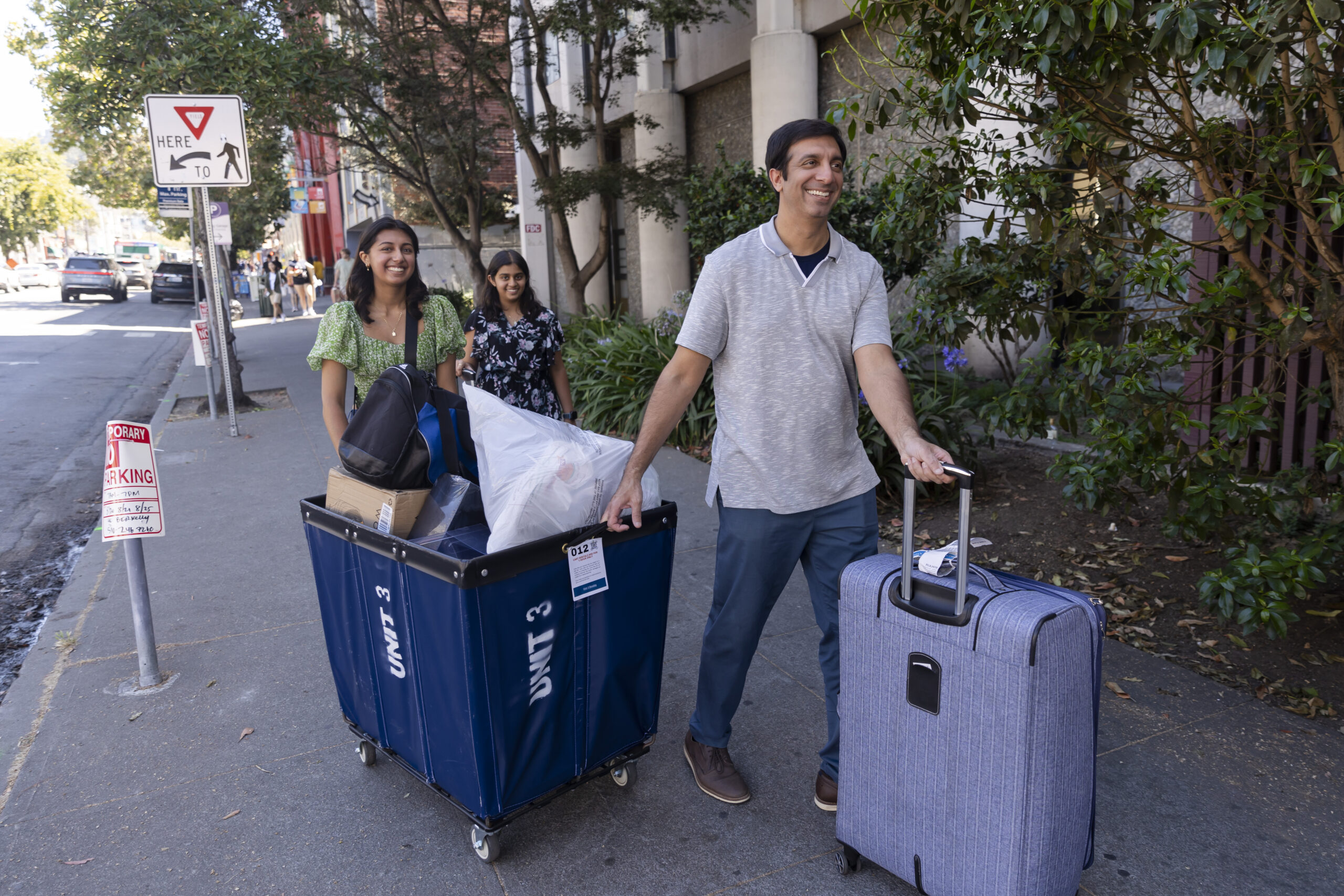 Families roll belongings into a residence hall using carts and luggage on wheels.