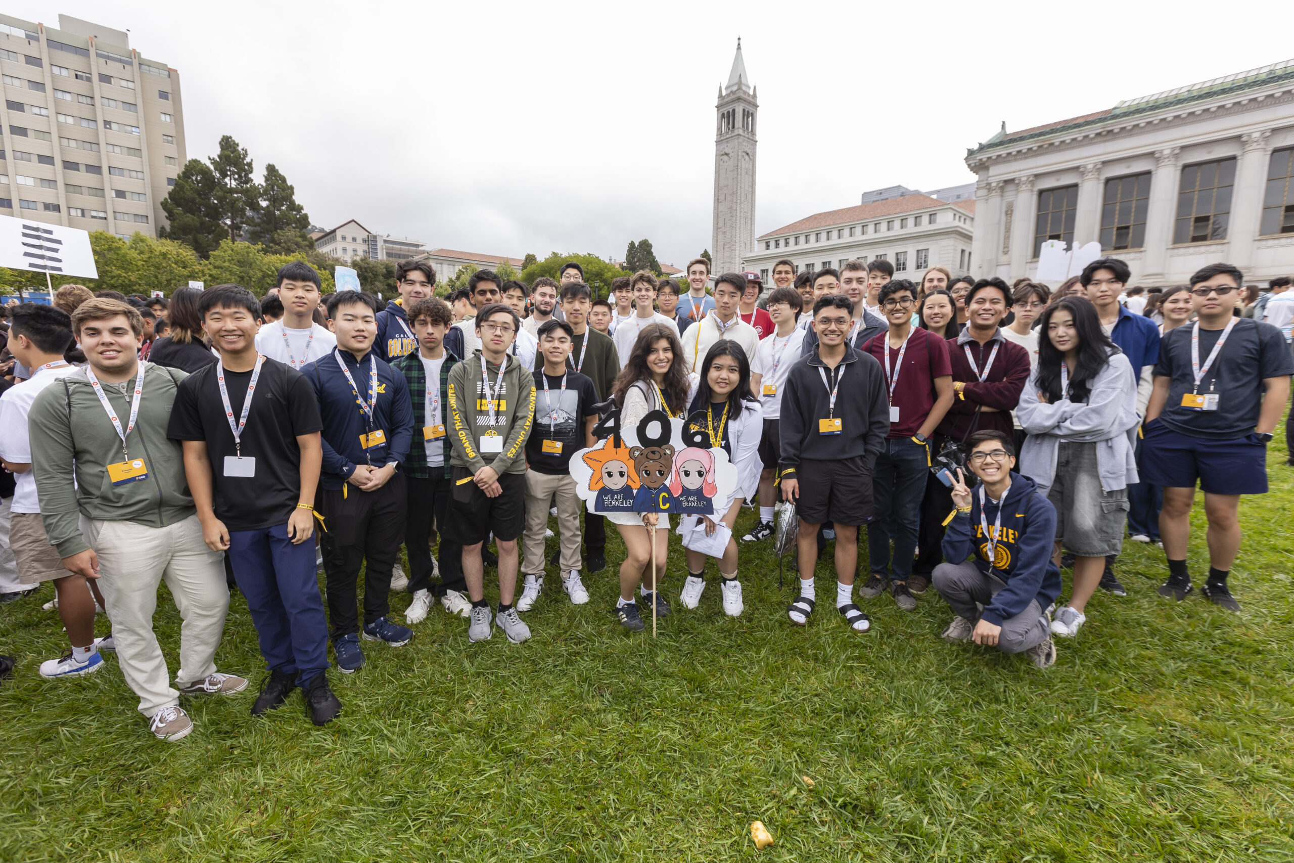 Dozens of students taking part in Golden Bear Orientation 2024 pose on Memorial Glade's green grass for a photo. They are in Group 406, one of many small groups of students that spend orientation together, with an orientation leader. The Campanile is in the background, and it's cloudy day.