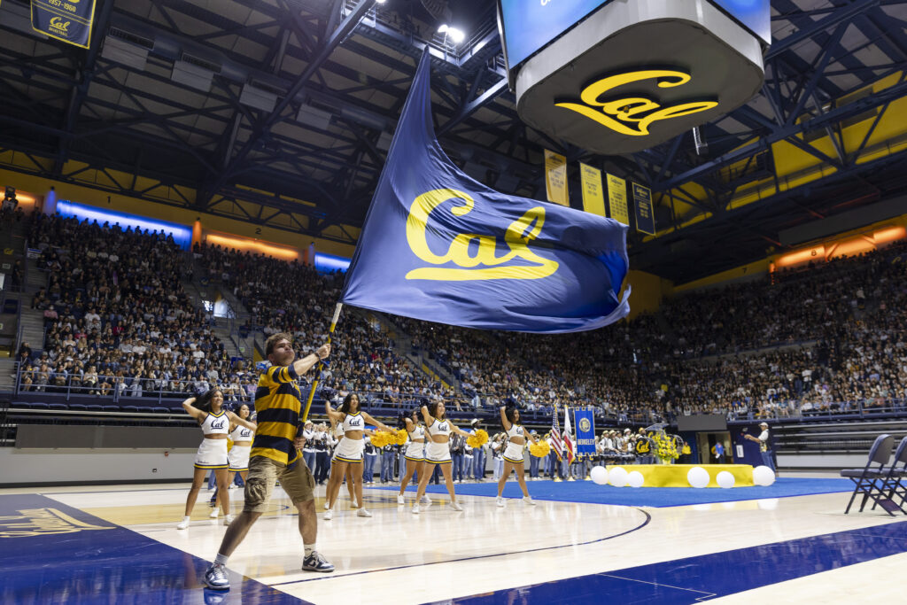 On the court of Haas Pavilion, members of Cal Cheerleading, the Cal Band and the Rally Committee perform during fall convocation. One student is waving a large Cal flag and the cheerleaders are holding gold pom poms. Students are sitting in the bleachers.