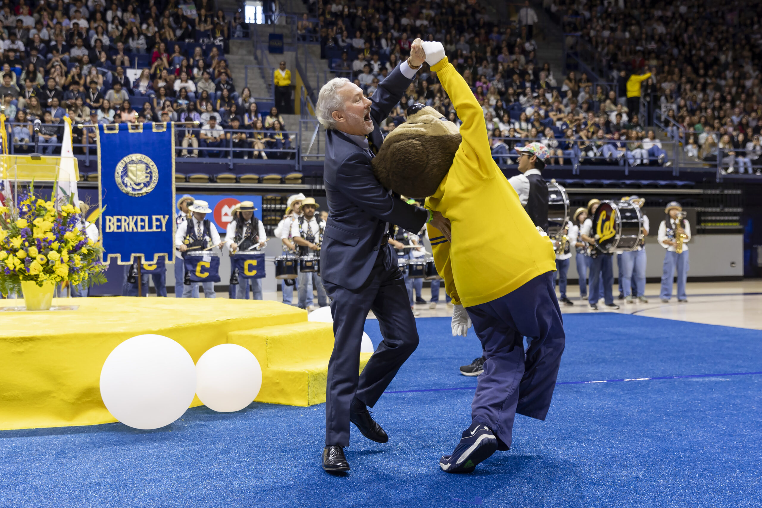 Rich Lyons, Berkeley's new chancellor, dances with Oski the Bear on the basketball court during fall convocation 2024. They are twirling each other around as members of the Cal Band play behind them.