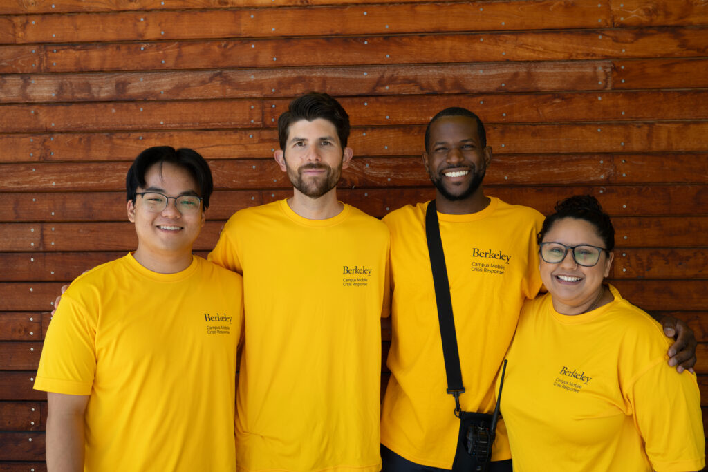 Four members of the Campus Mobile Crisis Response Program, all wearing gold T-shirts that say Berkeley Campus Mobile Crisis Response in dark blue letters on them, pose with their arms around each other in front of a building on campus with reddish-brown wooden siding.