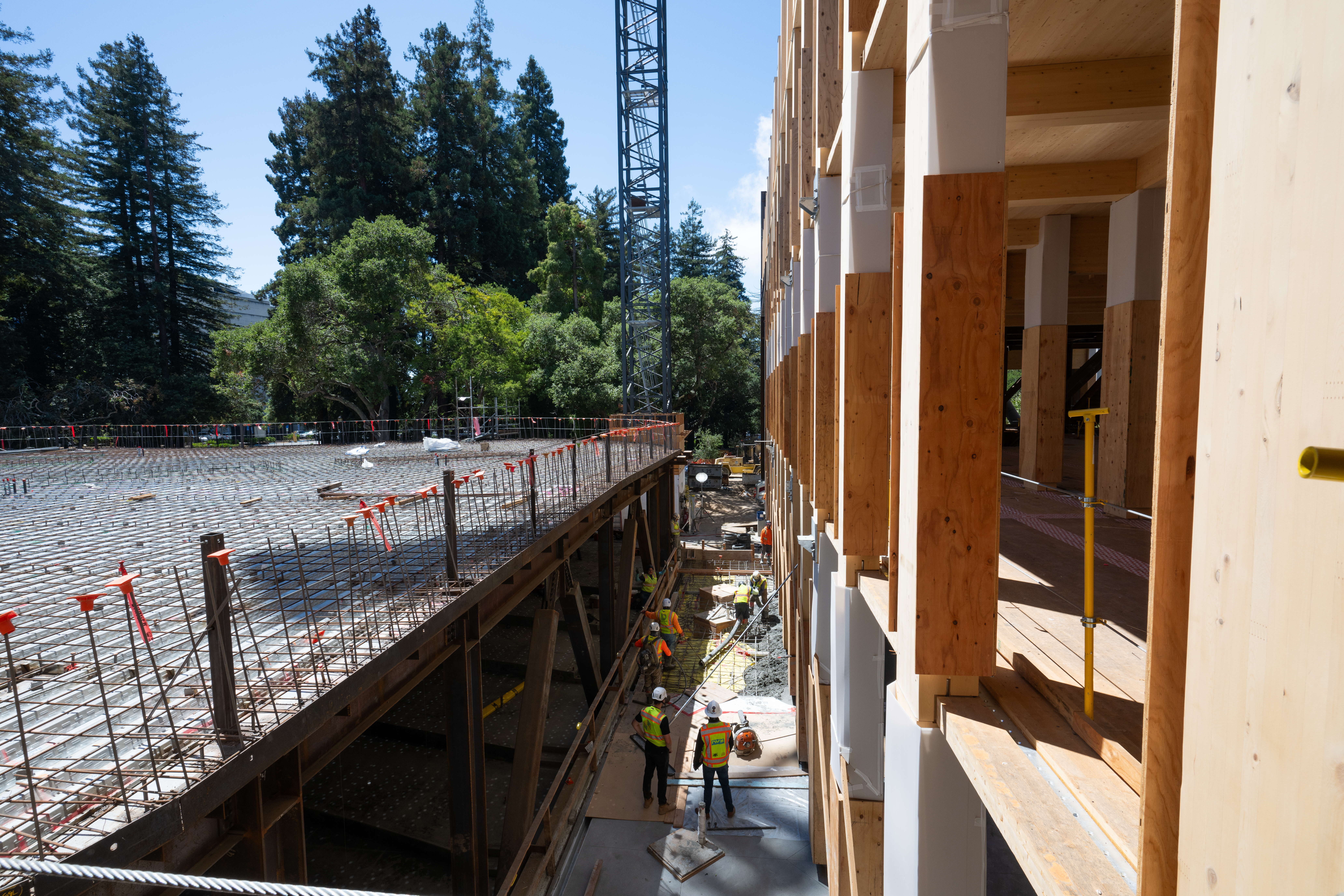 Workers in hard hats and security vests stand on the ground level of what will be the new Undergraduate Academic Building, in the center of campus. A large crane hovers above them.