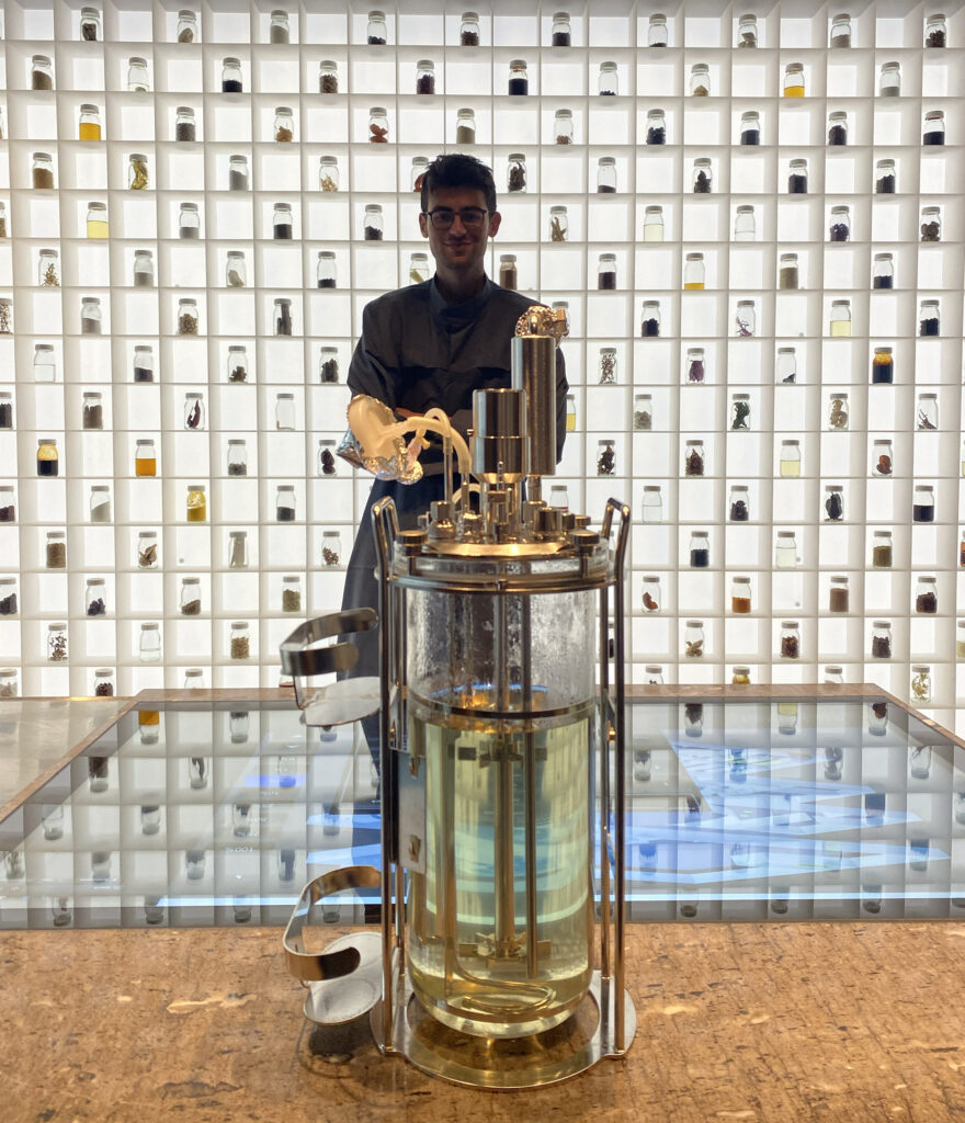 a dark-haired man in glasses stands next to a waist-high glass container of fluid and in front of a lighted wall of jars