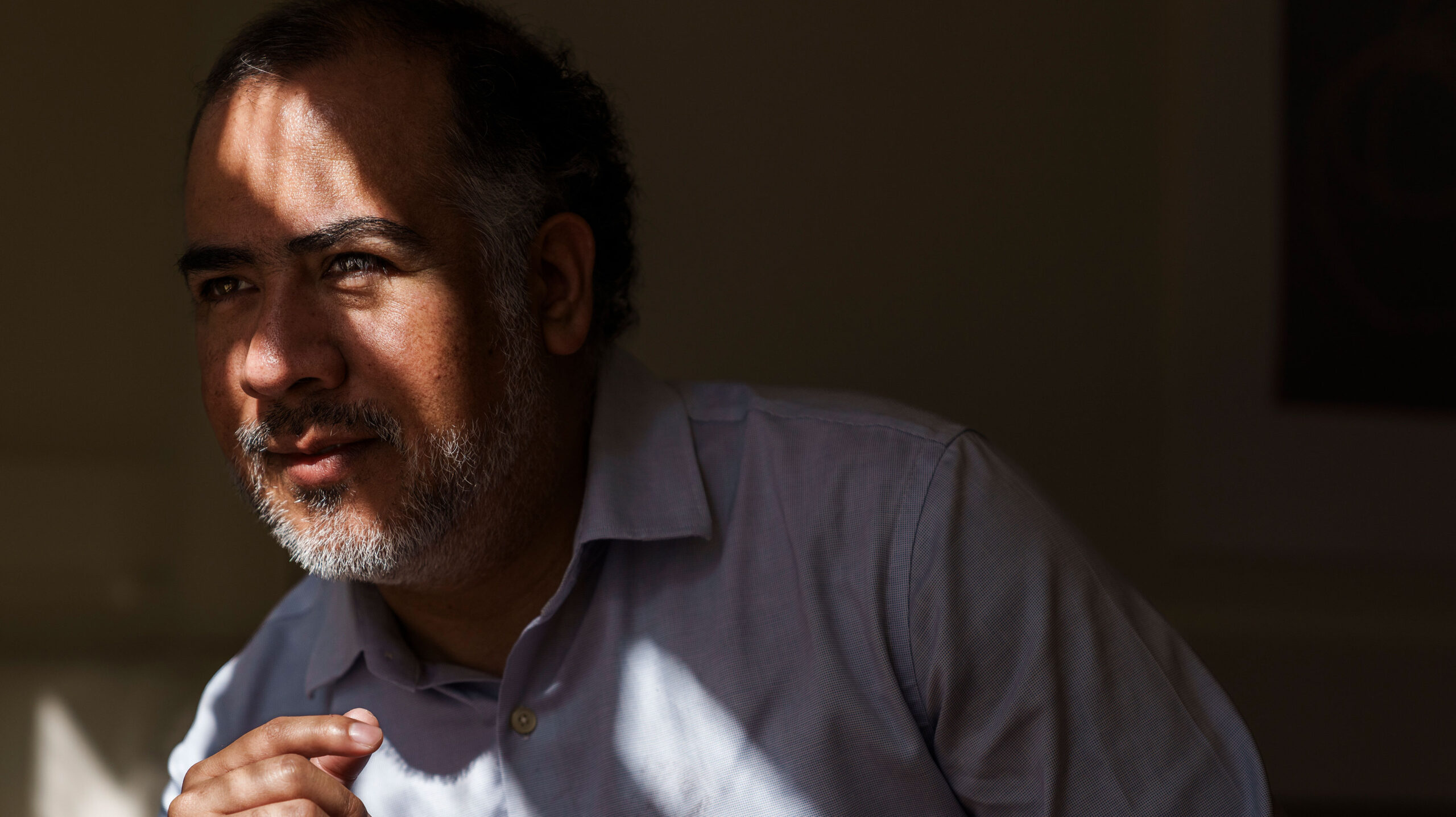 Haunting informal portrait of Omar Gómez Trejo, in deep shadows but with a few slashes of sunlight across his face and shoulders