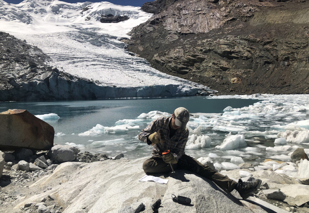 man in cap and gloves hammering on rock with glacier and glacial lake in the background