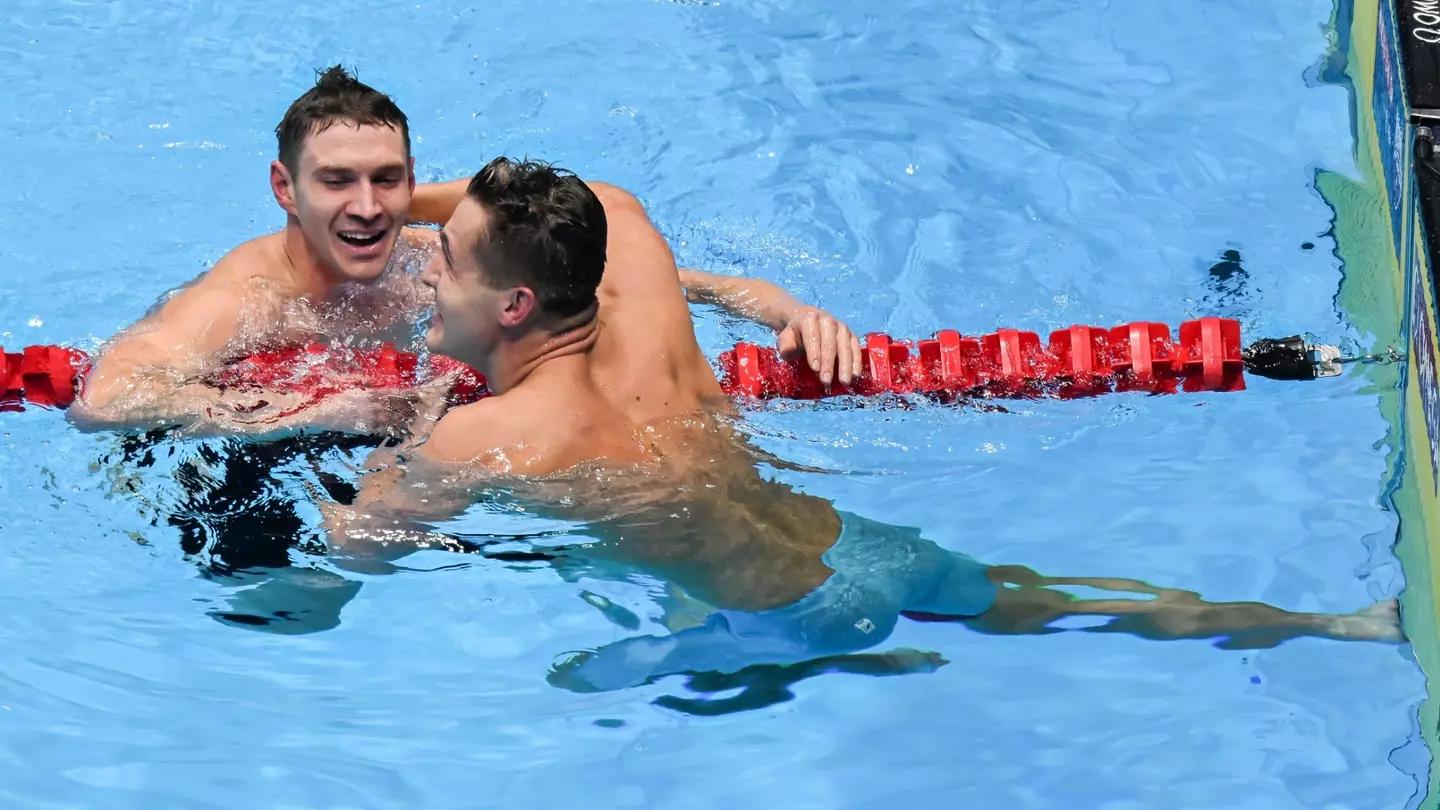 Two swimmers, both wearing blue swim trunks, celebrate in a swimming pool, smiling and embracing near a red lane divider. The water is clear and blue, with the swimmers showing signs of exertion and joy, possibly after a race or competition.