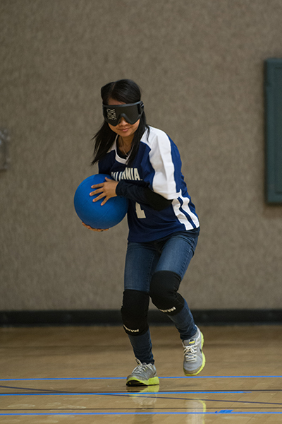 A student who is blind holds a ball and wears a mask over her eyes for the game of goalball, which she's playing at the Rec Sports Facility. The ball has bells inside and is playing by rolling the ball across the floor to the opponent's side of the court.