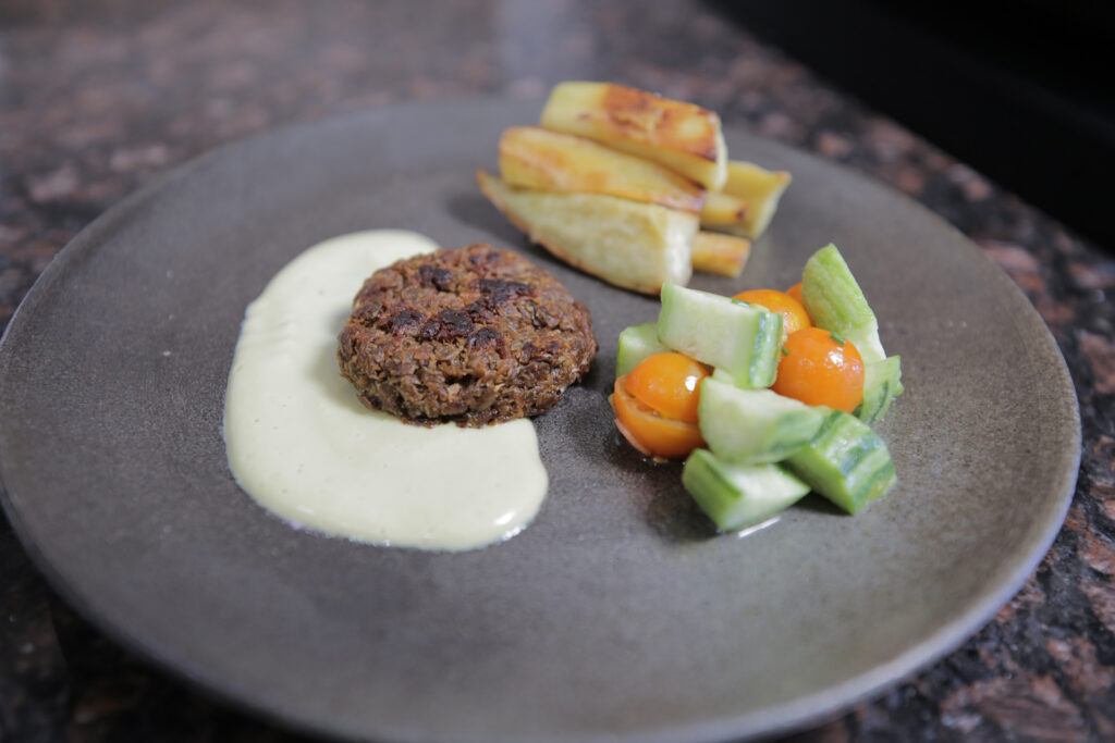 blue-gray plate with a seared patty sitting in a white sauce with roasted potatoes and cucumber tomato salad on side