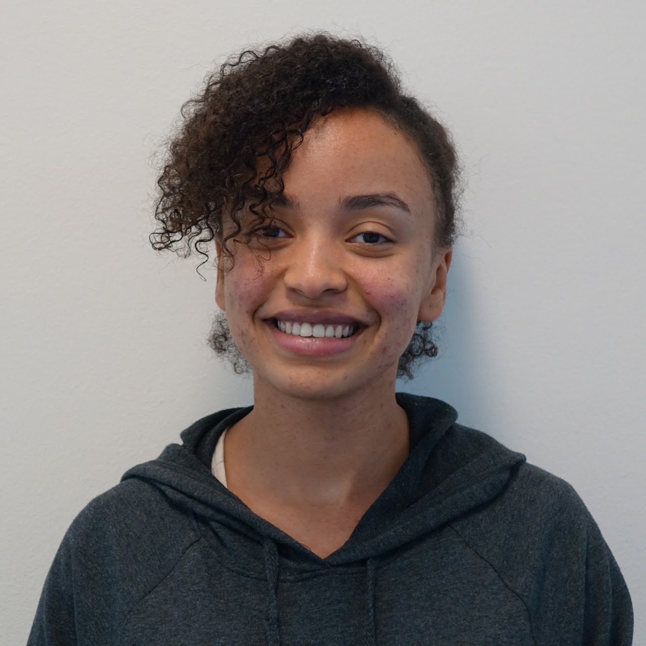 This image shows a person with curly hair and a friendly smile, wearing a dark hoodie, standing against a plain light-colored wall. The person's relaxed posture and cheerful expression convey a warm and approachable demeanor.