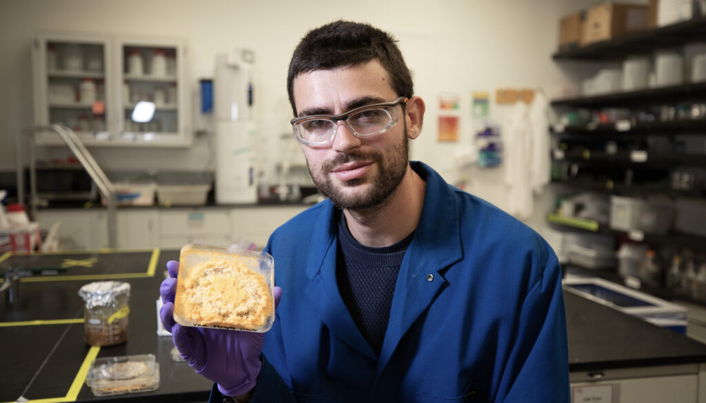 bearded man in dark blue lab coat holding a small tray filled with orange mold