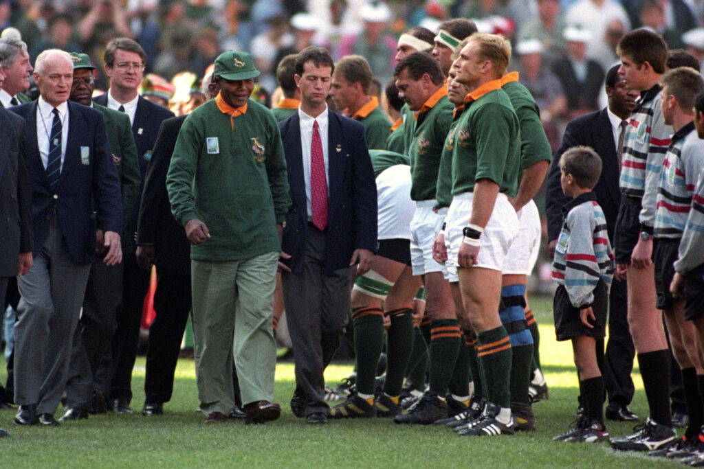 Nelson Mandela wearing a green Springboks cap and jacket as he walks onto a rugby field, alongside officials and players.