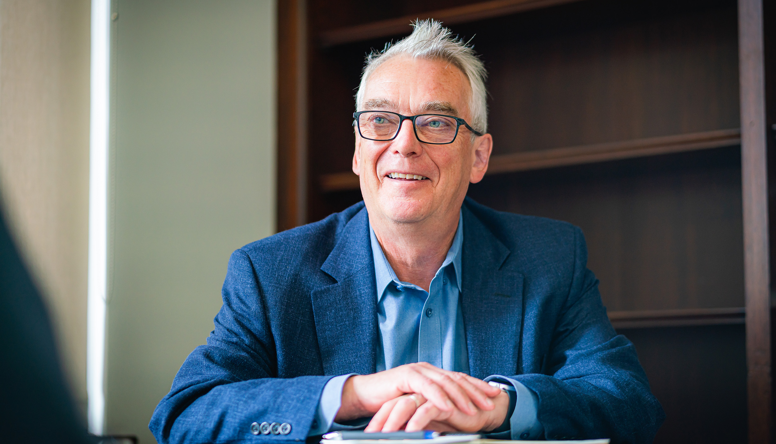 smiling man with glasses in a dark blue suit sitting at a desk