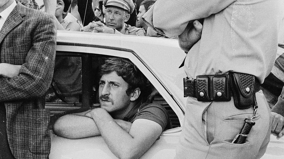 Jack Weinberg sits in a police car in 1964 with his arms folded out the window, surrounded by police and protesters