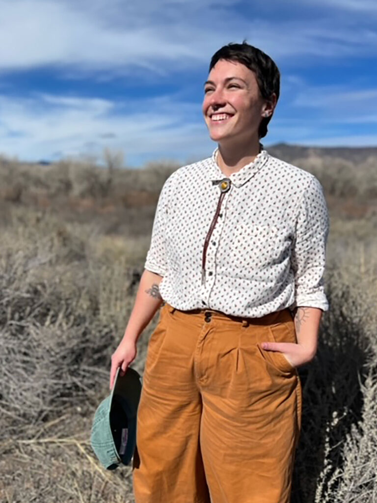 Julia McKeown, smiling broadly as they stand in a field of dry grass with a bright blue sky and wispy clouds in the background